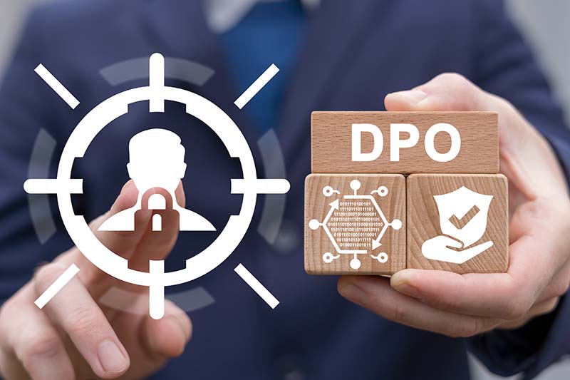 Une personne tient des cubes en bois avec les inscriptions "DPO" et des symboles de protection de données, sur fond d'icône cible de profil utilisateur et de cadenas, illustrant le rôle ciblé du Délégué à la Protection des Données.